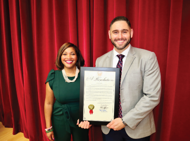 woman dressed in dark green standing next to man in suit holding an official letter